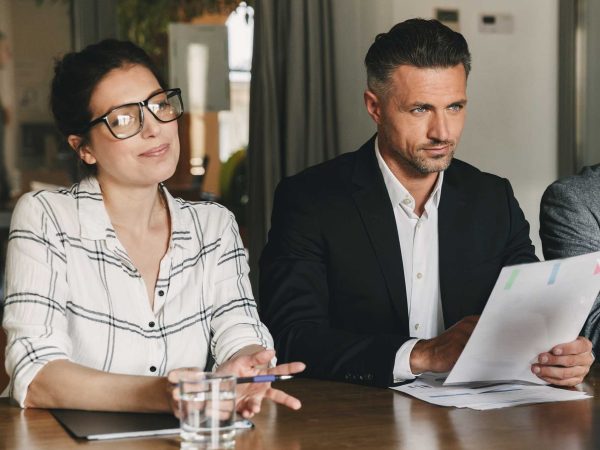Man and woman sitting at a table, woman holding a pen and man holding paper, both smiling.