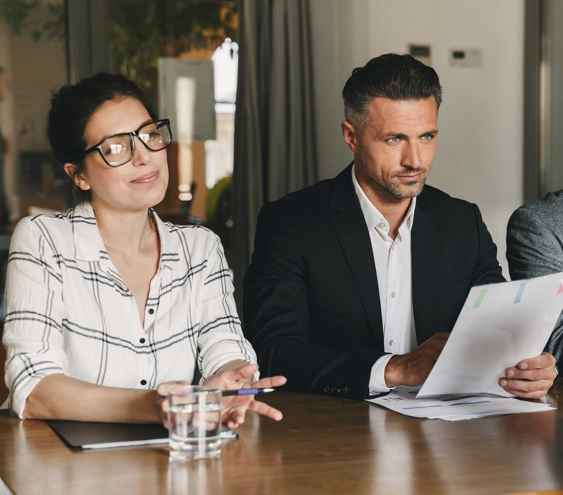 Man and woman sitting at a table, woman holding a pen and man holding paper, both smiling.