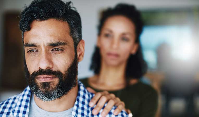 Grieving couple with man standing facing away and woman gently placing her hand on his shoulder."