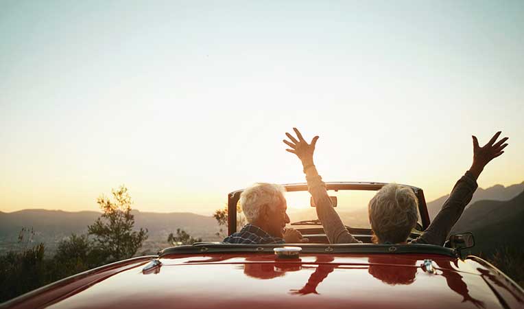 Retired couple in an open car with hay; man drives while woman raises her arms happily