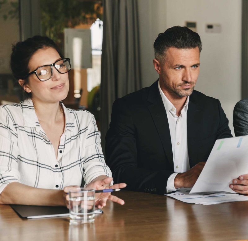 Man and woman sitting at a table, woman holding a pen and man holding paper, both smiling.